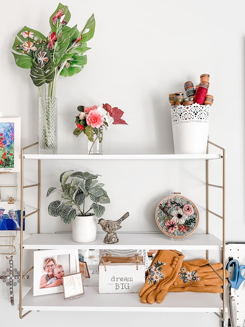 May include: A white shelf unit with gold metal accents, featuring a variety of decorative items, including a vase with pink and red flowers, a small potted plant, a bird figurine, a framed photo, a wooden box with the text "dream big", and a pair of embroidered gloves.