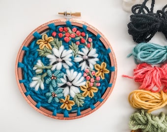 May include: A close-up of a finished embroidery hoop with a floral design. The hoop is made of wood and has a copper finish. The embroidery features white flowers, yellow flowers, pink flowers, and green leaves on a teal background. The embroidery is surrounded by a variety of colored embroidery floss.
