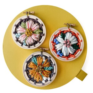 Pu&ograve; includere: Tre piccoli telai da ricamo rotondi con disegni floreali. Ogni telaio presenta una diversa combinazione di colori: rosa e verde, bianco e rosso e arancione e verde acqua. Il ricamo &egrave; posto su uno sfondo giallo.
