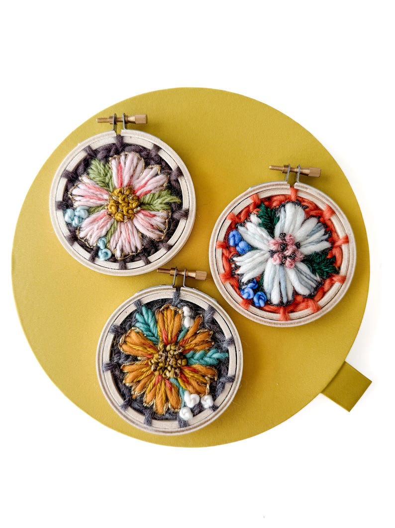 Pu&ograve; includere: Tre piccoli telai da ricamo rotondi con disegni floreali. Ogni telaio presenta una diversa combinazione di colori: rosa e verde, bianco e rosso e arancione e verde acqua. Il ricamo &egrave; posto su uno sfondo giallo.