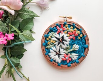 May include: A circular embroidery hoop with a blue background and colorful floral embroidery. The embroidery features white flowers with pink and yellow accents.