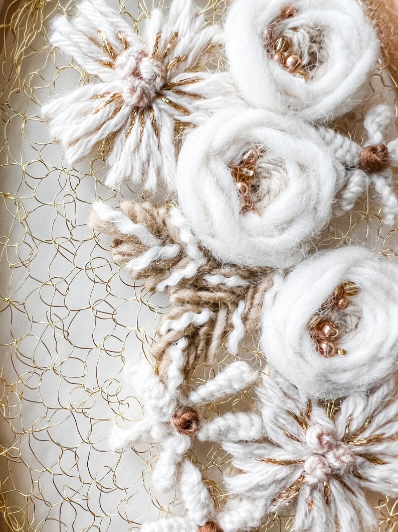 May include: A close-up of a white and gold floral embroidery on a gold wire mesh background. The embroidery features white yarn flowers with gold accents and brown yarn leaves.
