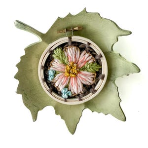 Pu&ograve; includere: Un telaio da ricamo fatto a mano con un fiore rosa e giallo, foglie verdi e dettagli blu. Il ricamo &egrave; posto in un telaio di legno e su una base di legno verde a forma di foglia.