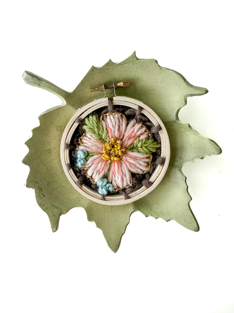 Pu&ograve; includere: Un telaio da ricamo fatto a mano con un fiore rosa e giallo, foglie verdi e dettagli blu. Il ricamo &egrave; posto in un telaio di legno e su una base di legno verde a forma di foglia.
