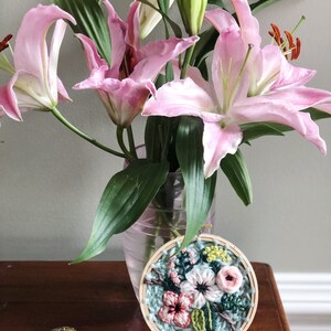 May include: A close-up of a small, round embroidery hoop with a floral design. The embroidery features pink, white, and green flowers on a blue background. The hoop is on a wooden stand and a gold key is in the foreground.