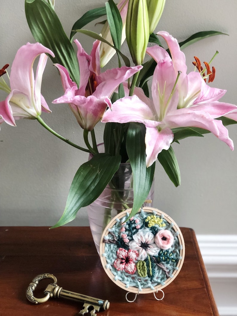 May include: A close-up of a small, round embroidery hoop with a floral design. The embroidery features pink, white, and green flowers on a blue background. The hoop is on a wooden stand and a gold key is in the foreground.