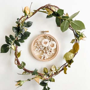 May include: A white floral embroidery hoop art piece with a brown wooden frame, surrounded by a wreath of green and brown vines with yellow flowers.