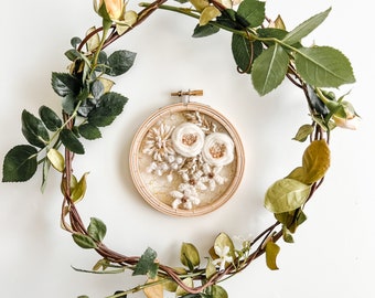 May include: A white floral embroidery hoop art piece with a brown wooden frame, surrounded by a wreath of green and brown vines with yellow flowers.