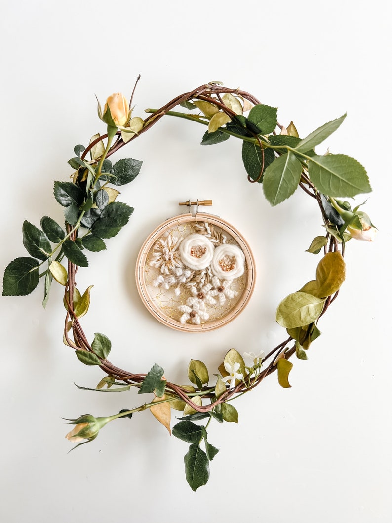 May include: A white floral embroidery hoop art piece with a brown wooden frame, surrounded by a wreath of green and brown vines with yellow flowers.