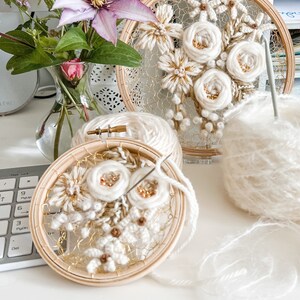 May include: Two embroidery hoops with white yarn flowers and gold accents. The hoops are on a white surface with a ball of white yarn.