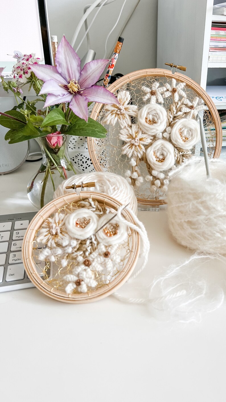 May include: Two embroidery hoops with white yarn flowers and gold accents. The hoops are on a white surface with a ball of white yarn.