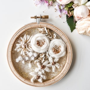 May include: A white and beige floral embroidery hoop art piece with gold accents. The hoop is made of wood and has a white background. The flowers are made of yarn and are arranged in a cluster. The piece is surrounded by a gold wire mesh.
