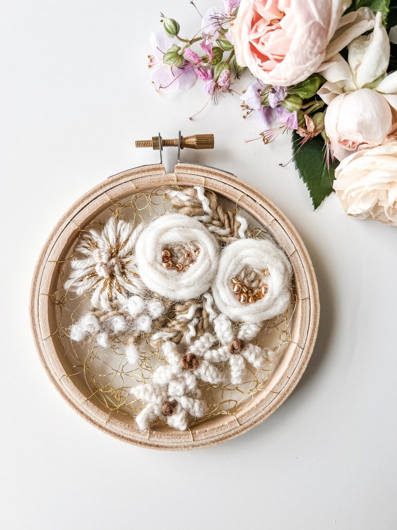 May include: A white and beige floral embroidery hoop art piece with gold accents. The hoop is made of wood and has a white background. The flowers are made of yarn and are arranged in a cluster. The piece is surrounded by a gold wire mesh.