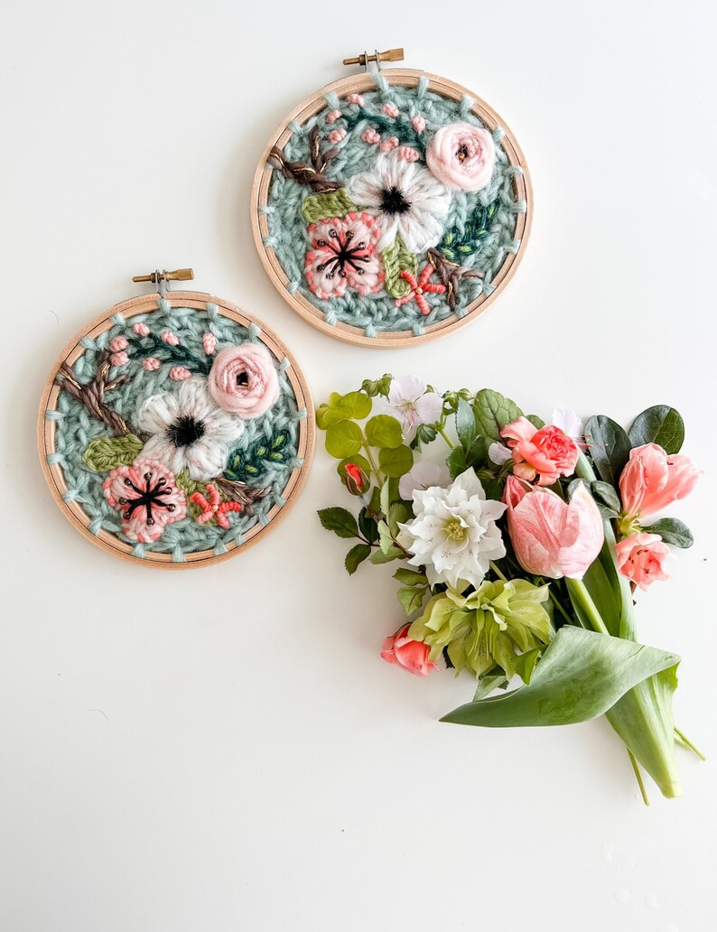 May include: Two embroidery hoops with floral designs. The hoops are made of wood and have a light green background. The flowers are pink, white, and green. A bouquet of pink and white flowers sits below the hoops.