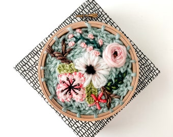 May include: A close-up of a floral embroidery hoop art piece. The hoop is made of wood and has a green fabric background. The embroidery features pink, white, and green flowers.
