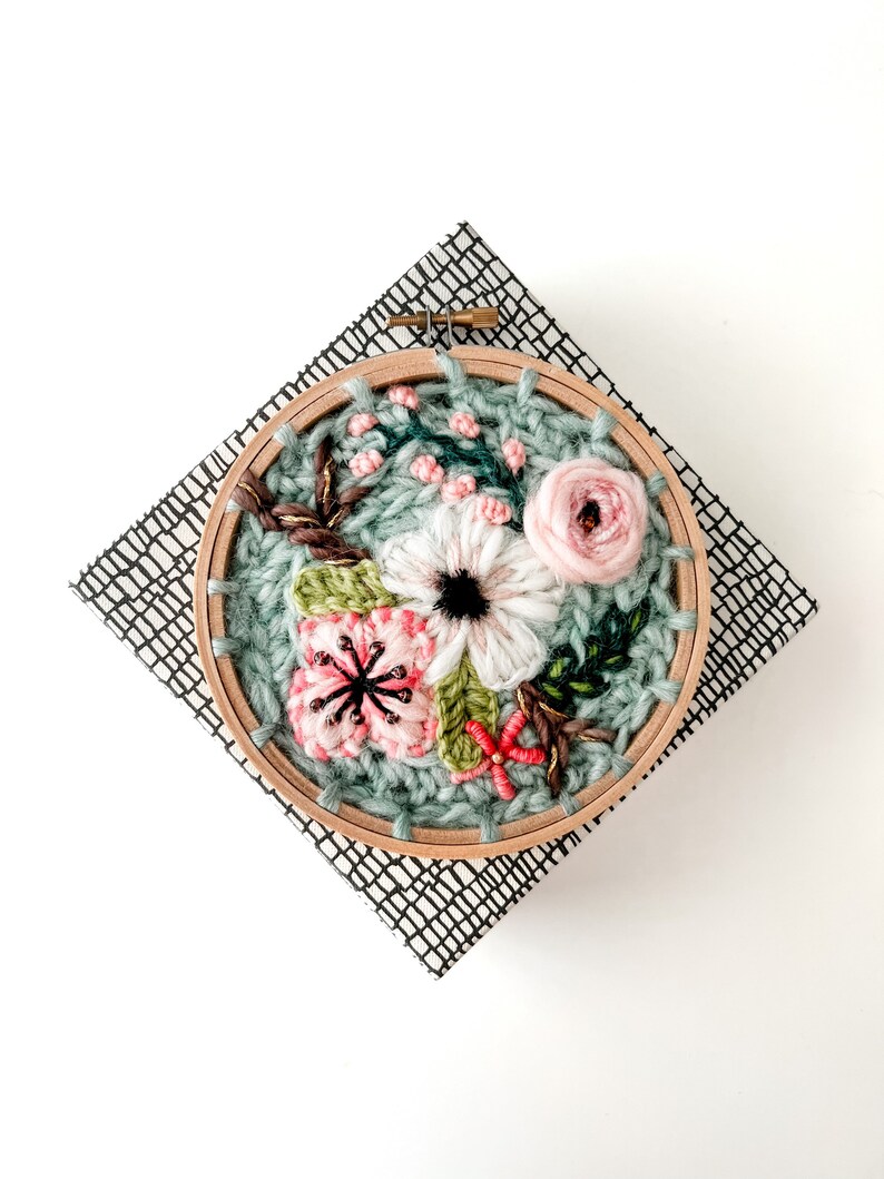 May include: A close-up of a floral embroidery hoop art piece. The hoop is made of wood and has a green fabric background. The embroidery features pink, white, and green flowers.