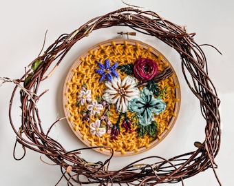May include: A circular embroidery hoop with a yellow background and colorful floral embroidery. The hoop is surrounded by a brown vine wreath.