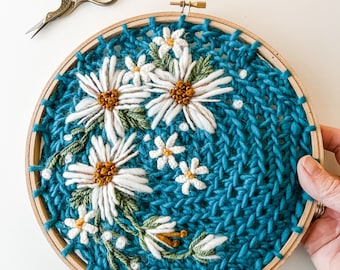 May include: A close-up of a circular embroidery hoop with a blue knitted background and white embroidered daisies with yellow centers. The daisies are arranged in a cluster and have green stems and leaves.