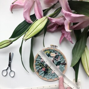 May include: A close-up of a hand holding a ruler measuring a circular embroidery hoop with a floral design. The hoop is made of wood and has a light blue background with pink and green flowers. The ruler is white with black markings and is 6 inches long. The hoop is surrounded by pink lily flowers and green leaves.