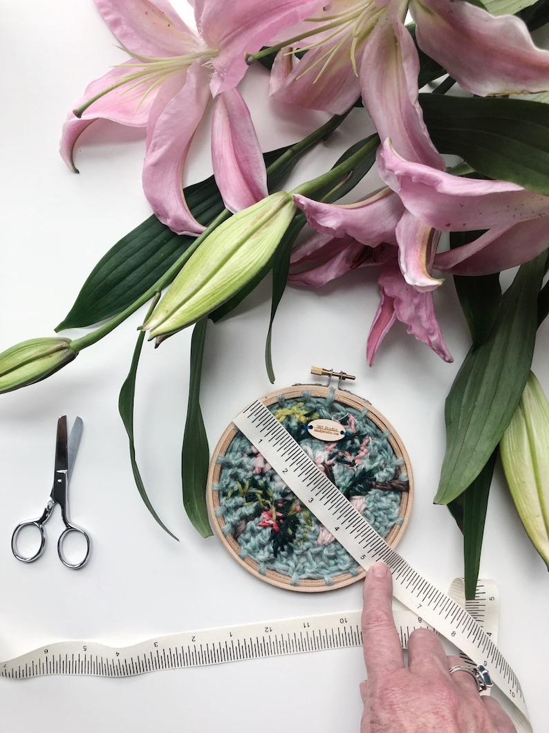 May include: A close-up of a hand holding a ruler measuring a circular embroidery hoop with a floral design. The hoop is made of wood and has a light blue background with pink and green flowers. The ruler is white with black markings and is 6 inches long. The hoop is surrounded by pink lily flowers and green leaves.