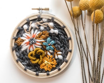 May include: A handmade fiber art piece in a wooden embroidery hoop. The artwork features a woven gray background with colorful embroidered flowers in white, orange, teal, and yellow. Dried yellow flowers are arranged to the right of the hoop.