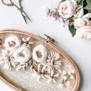 May include: A white floral embroidery hoop art piece with a gold wire mesh background. The hoop is made of wood and has a natural finish. The flowers are made of yarn and are arranged in a cluster. The embroidery is surrounded by a white background.