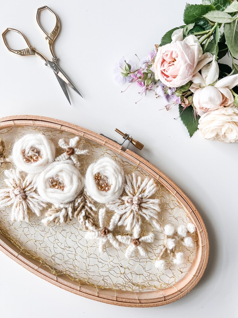 May include: A white floral embroidery hoop art piece with a gold wire mesh background. The hoop is made of wood and has a natural finish. The flowers are made of yarn and are arranged in a cluster. The embroidery is surrounded by a white background.