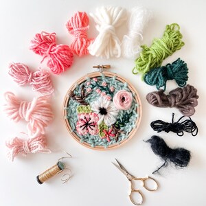 May include: A close-up of a floral embroidery hoop with a variety of colored embroidery floss, scissors, and a spool of gold thread. The embroidery hoop is filled with a colorful floral design.