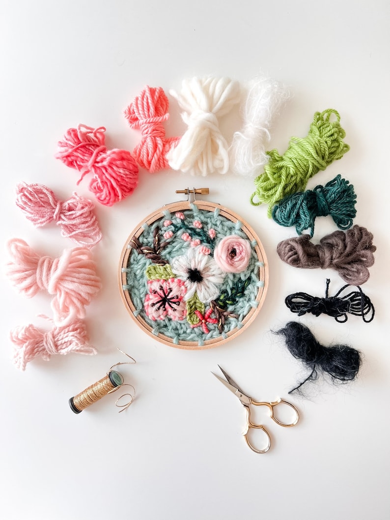 May include: A close-up of a floral embroidery hoop with a variety of colored embroidery floss, scissors, and a spool of gold thread. The embroidery hoop is filled with a colorful floral design.