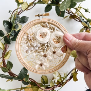 May include: A white floral embroidery hoop art piece with gold thread and beads. The hoop is surrounded by a wreath of green leaves and white flowers.