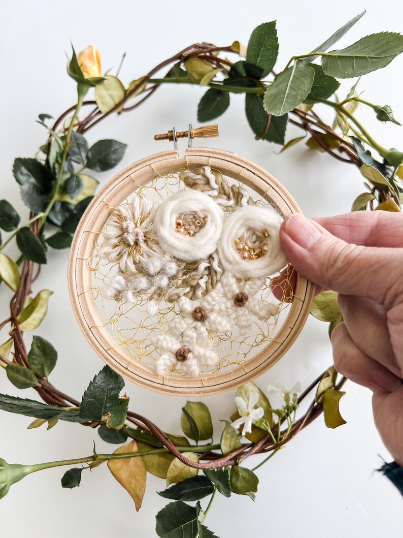 May include: A white floral embroidery hoop art piece with gold thread and beads. The hoop is surrounded by a wreath of green leaves and white flowers.
