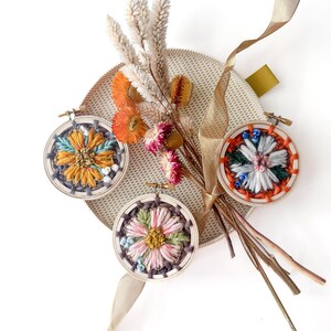 Pu&ograve; includere: Tre piccoli telai da ricamo rotondi con disegni floreali colorati. I telai sono disposti con fiori secchi e un nastro beige su una superficie circolare strutturata. Il ricamo presenta sfumature di giallo, blu, rosa e verde.