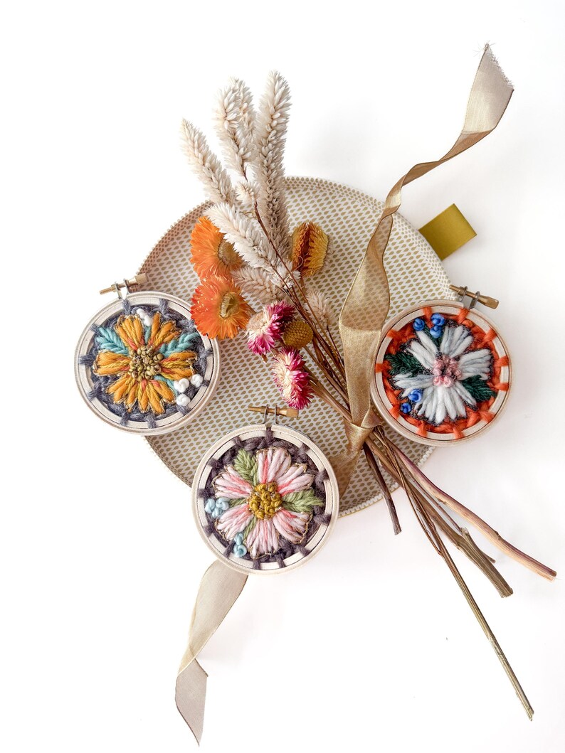 Pu&ograve; includere: Tre piccoli telai da ricamo rotondi con disegni floreali colorati. I telai sono disposti con fiori secchi e un nastro beige su una superficie circolare strutturata. Il ricamo presenta sfumature di giallo, blu, rosa e verde.