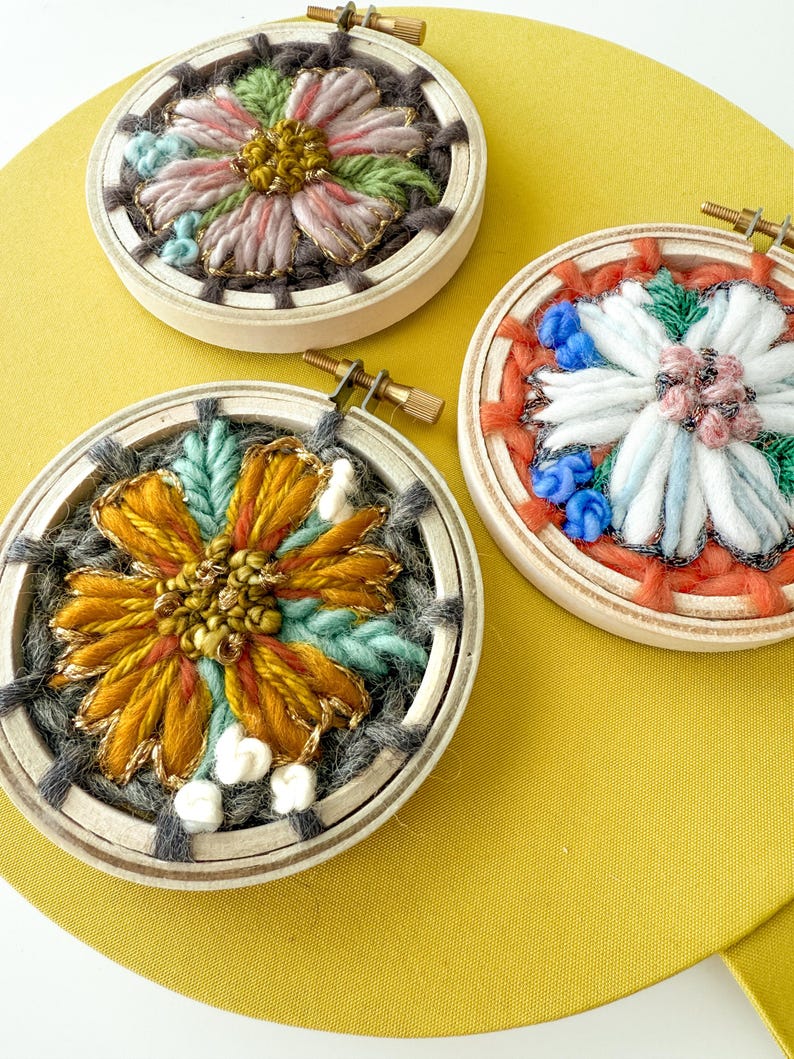 Pu&ograve; includere: Tre ricami floreali in cerchi di legno. I fiori presentano vari colori, tra cui rosa, arancione, giallo e blu, con accenti dorati. I cerchi sono posti su uno sfondo giallo.