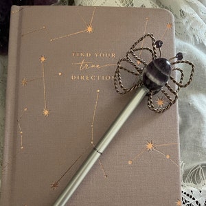 May include: A silver pen with a butterfly-shaped charm made of wire and a purple amethyst stone. The pen is resting on a closed journal with a constellation design and the text "Find your true direction".
