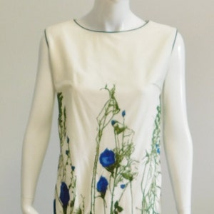 Könnte beinhalten: Ein weißes ärmelloses Kleid mit einem Blumendruck mit blauen Blumen und grünen Blättern. Das Kleid befindet sich auf einer Schaufensterpuppe und wird auf einer schwarz-weißen Polka-Dot-Oberfläche präsentiert.