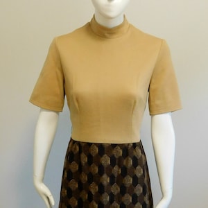 Op de afbeelding: Een beige jurk met een rok met geometrisch patroon. De jurk heeft een mock neck en korte mouwen. De jurk staat op een mannequin die op een zwart-witte stippen oppervlak staat.