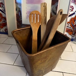 May include: A copper-colored utensil holder with a square shape, containing wooden kitchen tools. The tools include a slotted spoon, a spatula, and other wooden utensils. The holder sits on a white tiled surface.