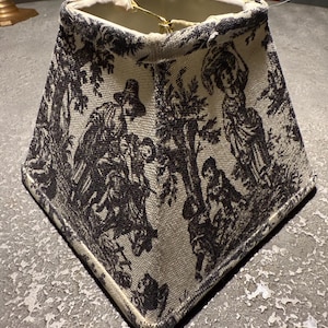 May include: A square lampshade with a black and white pastoral scene design. The fabric shade features a detailed print of figures in a landscape. The lampshade has a cream-coloured interior and a dark trim.