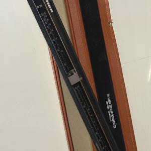 May include: A vintage black Gerber Scientific Instrument Co. slide rule in a brown leather case. The slide rule has a series of scales and markings, and the case is open, revealing the instrument. The text "THE GERBER SCIENTIFIC INSTRUMENT CO." is visible.