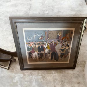 Könnte beinhalten: Gerahmter Kunstdruck, der eine Cafészene mit Figuren in verschiedenen Outfits darstellt. Das Kunstwerk zeigt ein Caféschild, Tische und Menschen, die sich unterhalten. Der Rahmen hat ein dunkles, abgewinkeltes Design.