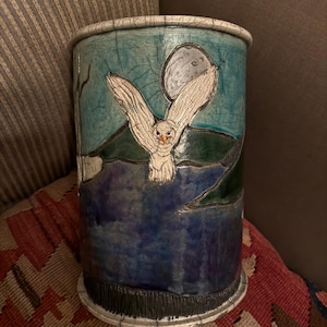 May include: A cylindrical ceramic vase with a hand-painted owl design. The owl is depicted in flight against a night sky with a full moon. The vase features shades of blue, green, and white, with a textured finish. The base and rim have a dark, textured border.