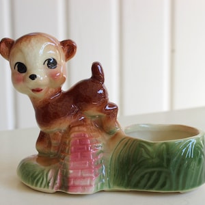 May include: A ceramic planter shaped like a deer with brown and white fur, standing on a pink bridge with green foliage. The planter has a small, round opening for planting.