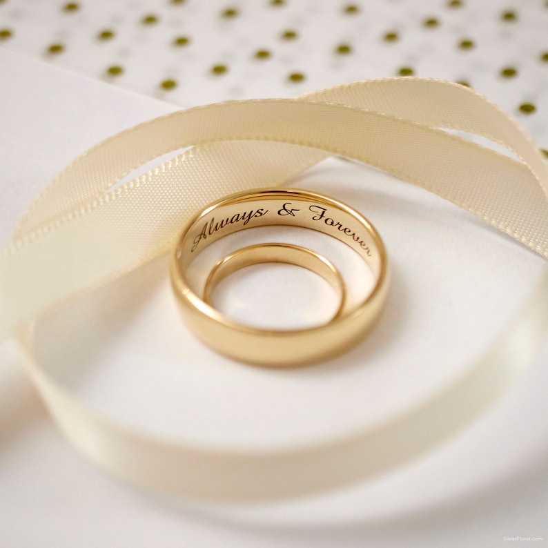 May include: Two gold wedding bands with the inscription "Always & Forever" on the outside of the larger band. The bands are on a white background with a cream-colored ribbon.