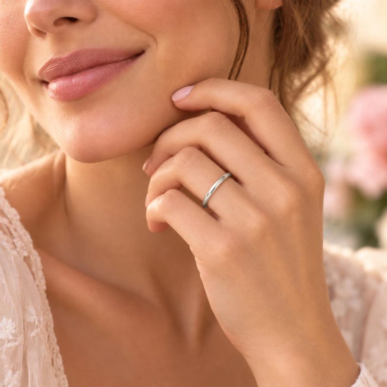 May include: A simple, polished silver-toned band ring with a smooth, rounded surface. The ring features a minimalist design with a high-shine metallic finish, worn on the ring finger of a hand, showcasing its clean, reflective, and circular form.