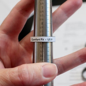 May include: A ring sizer tool with a ring marked "Comfort Fit - US 9" is being used to measure a ring size. The tool is silver and has measurements in inches. The background includes other rings and a sheet of paper.