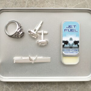 May include: A silver tray with a silver ring, cufflinks, a tie bar, and a small tin of solid cologne. The cologne tin has a blue and white label with the words "Jet Fuel" and a picture of a fighter jet. The label also says "Solid Cologne" and "TheSouthernWolf.com".