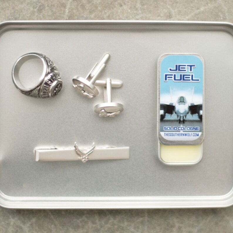 May include: A silver tray with a silver ring, cufflinks, a tie bar, and a small tin of solid cologne. The cologne tin has a blue and white label with the words "Jet Fuel" and a picture of a fighter jet. The label also says "Solid Cologne" and "TheSouthernWolf.com".