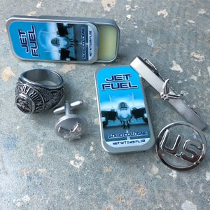 May include: A silver ring with a detailed design, a pair of silver cufflinks with a bird design, a silver tie clip with a bird design, a silver US charm, and two silver tins of Jet Fuel solid cologne. The cologne tins feature a blue background with a fighter jet design and the text "Jet Fuel Solid Cologne" and "Net Wt 0.25 fl oz".
