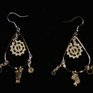 May include: A pair of silver earrings with a teardrop shape. Each earring has a small gear charm, a small key charm, and a small metal charm. The earrings are hanging from silver hooks.